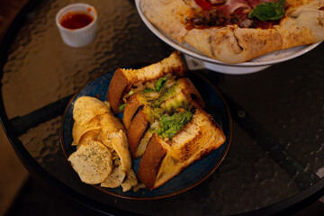 Prawn toast sandwich, stacked on a black plate, served alongside potato chips, in a cafe environment