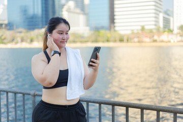 Fototapeta premium Portrait of young person in sportswear checks their phone by a waterfront, surrounded by urban buildings, suggesting an active lifestyle and modern environment. Asian plus size woman