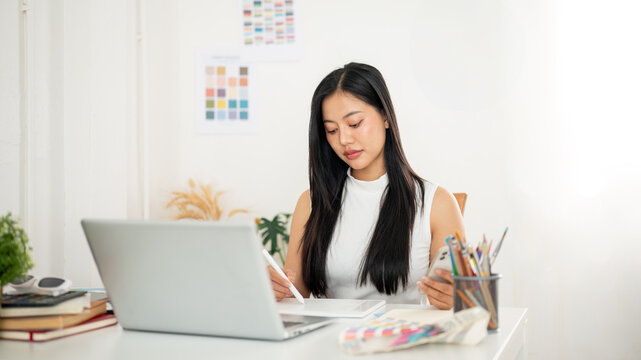 An asian woman is holding a stylus drawing or writing on graphic tablet while holding a smartphone.