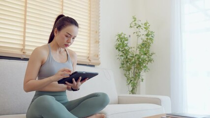 A woman is sitting on a couch and using a tablet. She is wearing a grey tank top and green pants