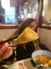A young girl in a restaurant holds a freshly made spring roll with chopsticks, offering a close-up view of the food.