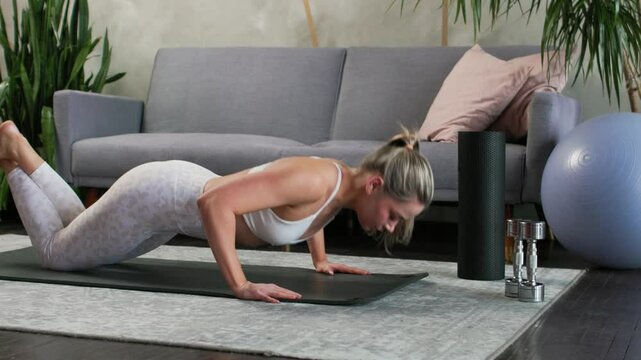 Young beautiful blonde doing knee push-ups on yoga mat in cozy living room. Gray sofa and houseplants, palm trees, dracaenas in the background. Active lifestyle concept.