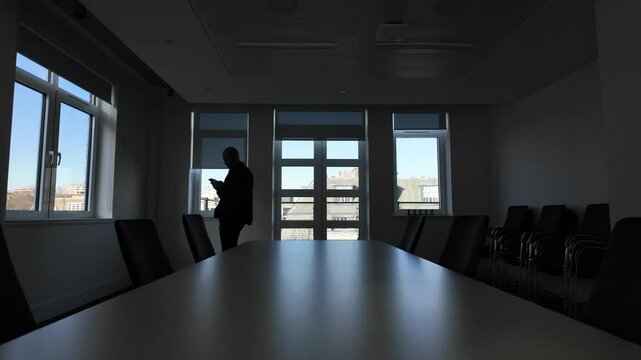 Silhouette of a businessman walking through a meeting room, engaged with a smartphone, surrounded by modern office furniture and shadows