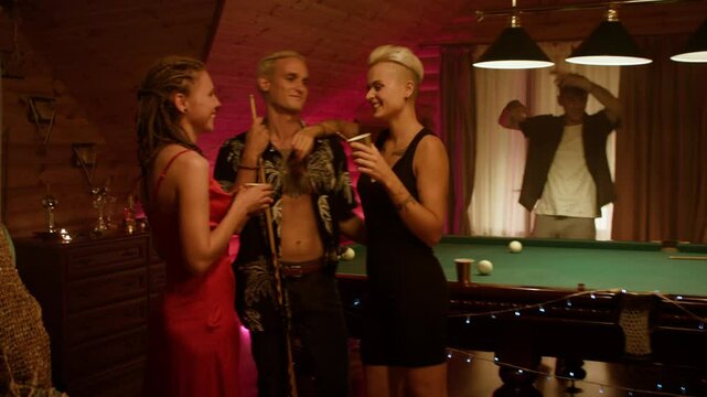 Cozy wooden party: a woman in a black dress with short light hair embraces a smiling man, hand on shoulder and cup in hand, while a red-dressed friend with dreads smiles by the billiard table.
