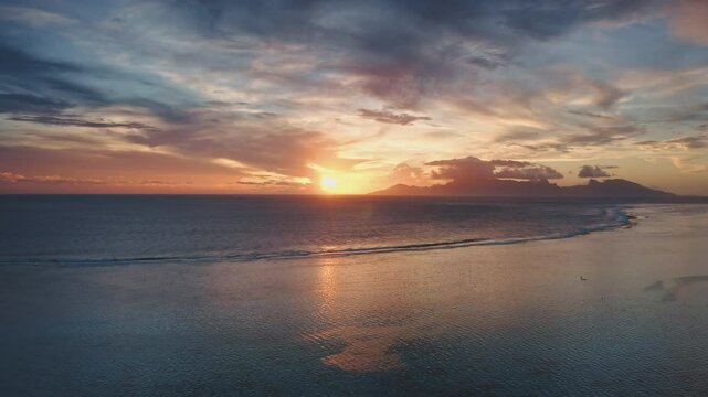 Golden sunset illuminating the calm ocean waters surrounding tropical Tahiti island with volcanic mountains visible on the horizon under a cloudy sky