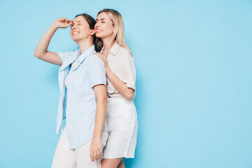 Naklejka premium Two young beautiful smiling brunette and blond hipster female in trendy summer clothes. Lovely women posing on blue background in studio. Positive models having fun. Cheerful and happy. isolated