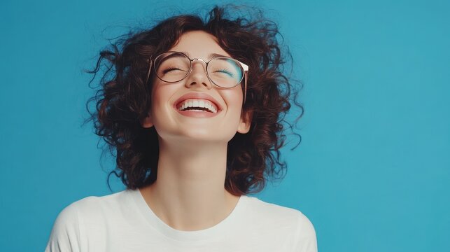 Waist up shot of happy curly woman with toothy smile, wears optical glasses and casual solid white t shirt, expresses good emotions, enjoys nice day, isolated over blue background. Face expressions