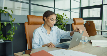 smiling young woman in glasses sitting at a modern office desk, closing her laptop while enjoying a...