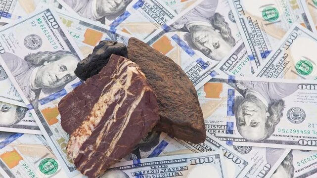 Three gemstones, Tiger Iron, Hematite, and Obsidian, rotate on a turntable atop genuine US 100-dollar bills. Dark stones with metallic and earthy tones contrast with the green currency background.