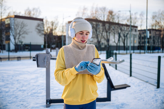 woman with headphones using smartphone in snowy outdoor gym