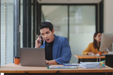 Young entrepreneur managing his business, answering phone calls and using laptop, sitting at desk...