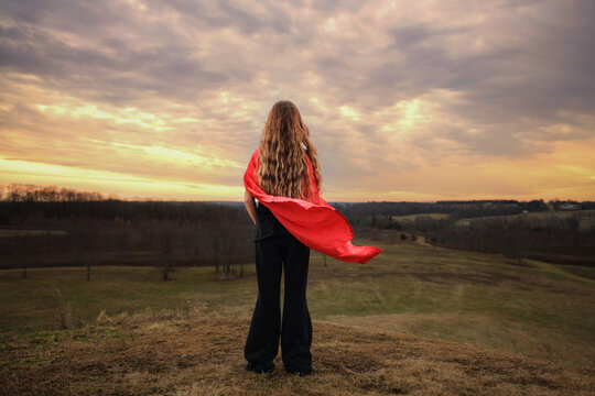 Behind young girl standing on hill red cape blowing dramatic sky