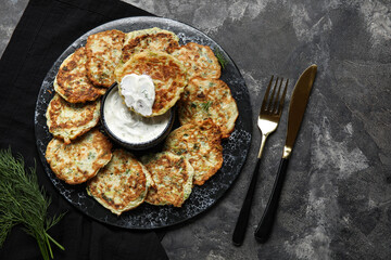 Board of tasty zucchini fritters with sour cream on dark background