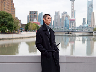 Portrait of handsome Chinese young man wearing black overcoat posing in the street, young guy with black short hair with Shanghai bund background. Male fashion, cool Asian young man lifestyle.