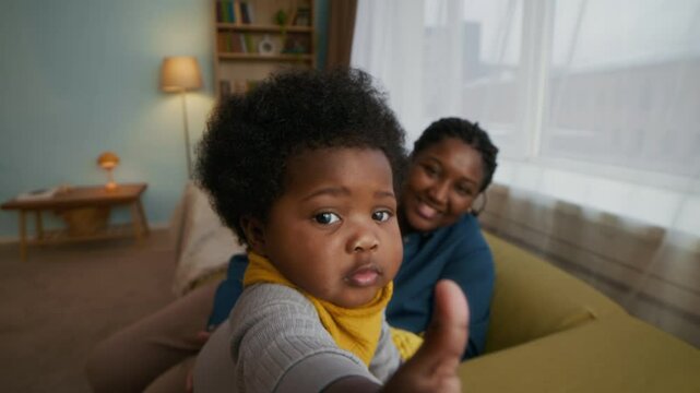 Portrait of curious African American toddler in yellow bib trying to grab camera from couch, while her mother holding her and smiling