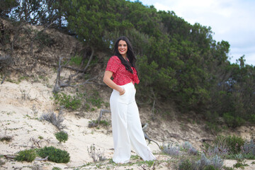 Young, brunette woman barefoot in the dunes by the beach. The woman is smiling and happy and has her hands in her pocket. The woman is wearing white trousers and a red shirt.