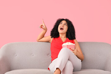 Beautiful woman with popcorn on sofa pointing at something against pink background