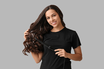 Beautiful young woman with curling iron on grey background