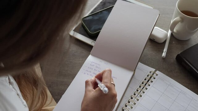 Top-down view of woman writing tasks in to-do list while sitting at desk with planner, coffee, and digital devices. Productivity and organization theme.