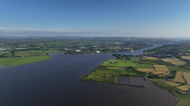 Lough Foyle, County Donegal, Ireland, June 2023. Drone pulls backwards in a panoramic high-angle view above the water, with rural fields and farms below rolling hills on the horizon under a clear sky.