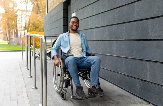 Handicapped accessible city concept. Positive impaired Afro man in wheelchair leaving building on ramp outdoors in autumn, full length. Joyful young black guy using disabled friendly facilities