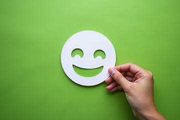 Hand holding a white paper smiley face on a vibrant green background, symbolizing happiness, positivity, and joy in a simple, minimalist design