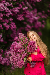 Naklejka premium Cute little girl in pink coat holds big bouquet of lilacs in her hands and smiles in spring park near lilacs 