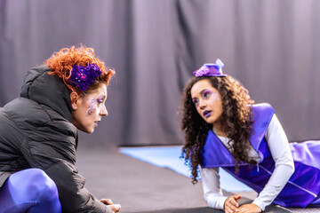 Two actresses wearing colorful costumes and makeup are rehearsing a scene for a futuristic play on a stage