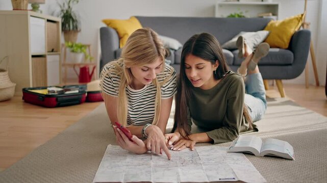Young female friends planning a trip together at home using cell phone travel app and a guidebook. Pointing at city map
