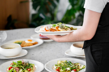 A waiter expertly carries a plate of gourmet dishes in a modern restaurant setting, surrounded by beautifully arranged plates on a gray stone table.