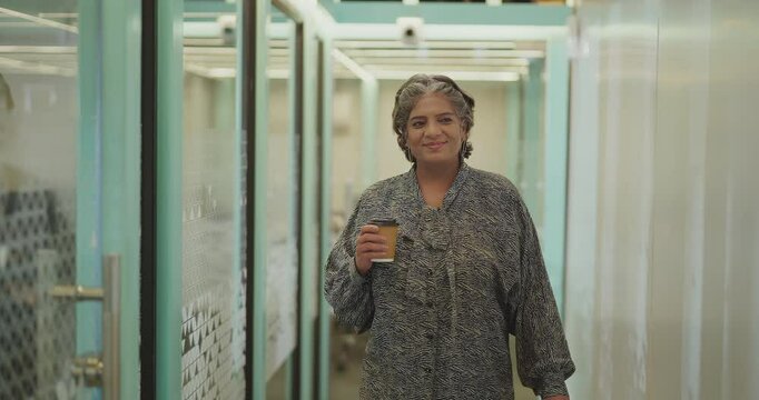 Indian gen x happy senior old aged boss woman lady ceo hold paper cup walk indoor office space smiling elder older active female staff worker drink coffee spend day time break hall way job work place