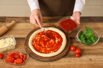 Woman spreading tomato paste on dough for pizza near table in kitchen