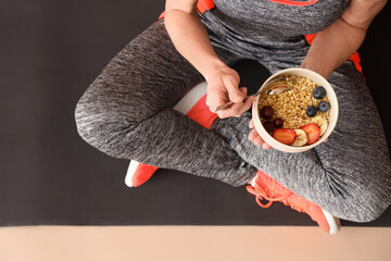 Mature sporty woman with bowl of nutritious oatmeal and fruits sitting on yoga mat, closeup