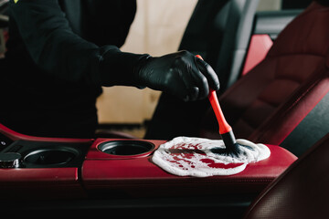 Washer applies foam to the car's red leather for interior cleaning at the service. Car detailing process of cleaning leather auto interior with soap foam at the carwash.