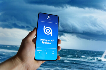 Hand holding a smartphone displaying a weather app with a hurricane or typhoon warning. Dark storm...