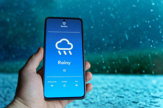 Hand holding a smartphone displaying a weather app with a rainy forecast. Raindrops falling in the blurred background. Weather technology and forecast concept