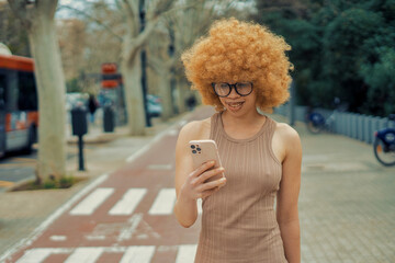 Stylish urban girl with glasses and bright orange afro hair is using her smartphone while walking along a city bike lane