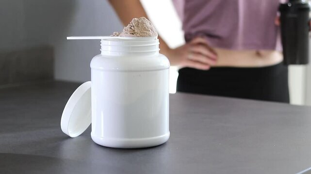 Close up of whey protein powder box and pink dumbbells on table, woman preparing a protein shake
