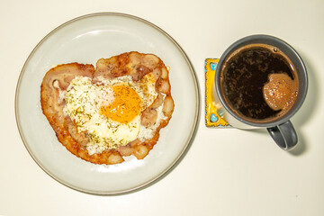 Delicious breakfast with a fried egg on crispy bacon and a cup of black coffee with a heart-shaped foam. A cozy morning meal served on a white plate with a decorative coaster