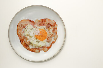 A top-down view of a breakfast dish featuring a fried egg with a runny yolk on crispy bacon, garnished with herbs and grated cheese, served on a white plate. A delicious and hearty morning meal