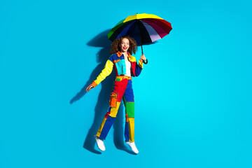 Cheerful young woman in vibrant colorful outfit holding rainbow umbrella against bright blue...
