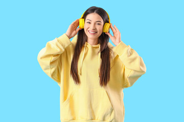 Stylish young woman in yellow hoodie with headphones on blue background