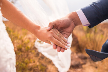 Noivo e noiva de mãos dadas com aliança de casamento e vestido na sessão pós wedding externa. 