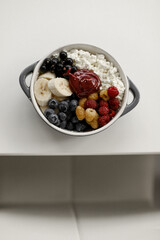 A plate with cottage cheese, blueberries, red and yellow raspberries, banana pieces, and berry honey stands on a white table