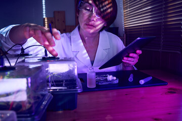Female scientist with digital tablet examining plants growing in greenhouse farm with neon phyto...
