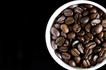 A top-down view of glossy roasted coffee beans in a white ceramic mug on a deep black background, perfect for coffee ads, menus, or branding. Photo. Copy space