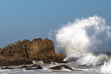 Giant wave breaking against large rock just offshore Malibu, California. Spray flying high in the air; blue sky in the distance. 
