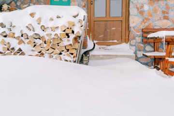 Tabby cat stands near a woodpile on the snow-covered threshold of a stone house near the door