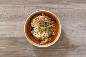 Overhead shot of a gourmet meal served in an eco-friendly take-away bowl, featuring stuffed eggplant with seasoned meat, sitting in a rich tomato sauce. Placed on a wooden surface with copy-space