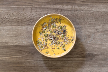 Creamy pumpkin soup topped with mixed seeds, served in an eco-friendly bowl. A warm and healthy option for a restaurant menu or take-away service. Plated on a wooden surface